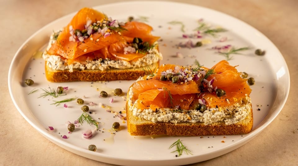 Two pieces of toast with smoked salmon, cream cheese, capers, and red onion on a white plate.