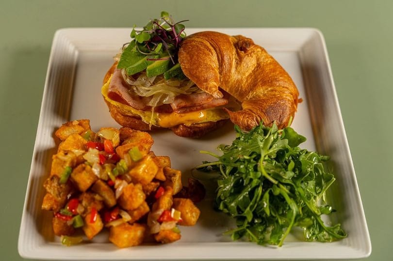Golden croissant sandwich with ham, egg, cheese, avocado, potatoes, and fresh arugula on a white plate.
