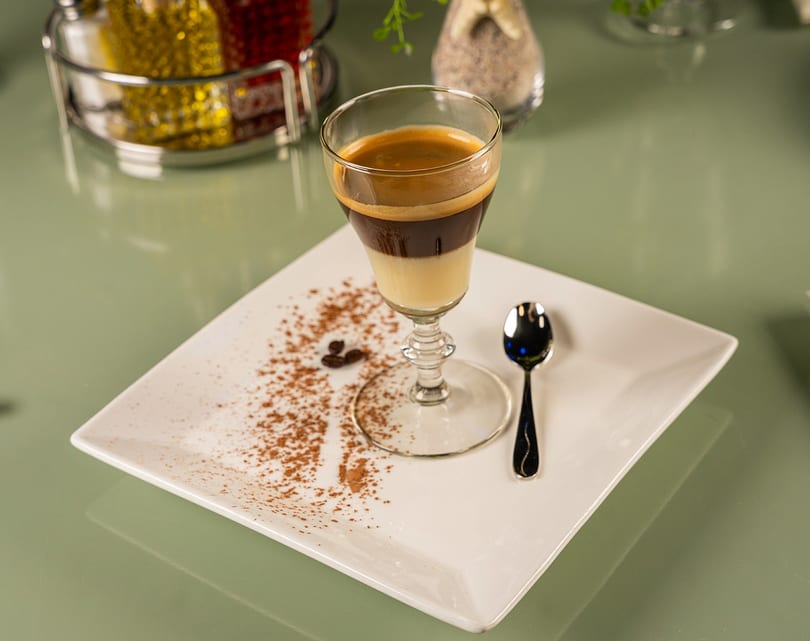 Layered coffee drink in a stemmed glass on a white plate with cocoa powder and a spoon.