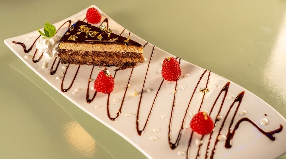 A slice of chocolate cake with gold flakes, whipped cream, mint, and raspberries on a white plate.