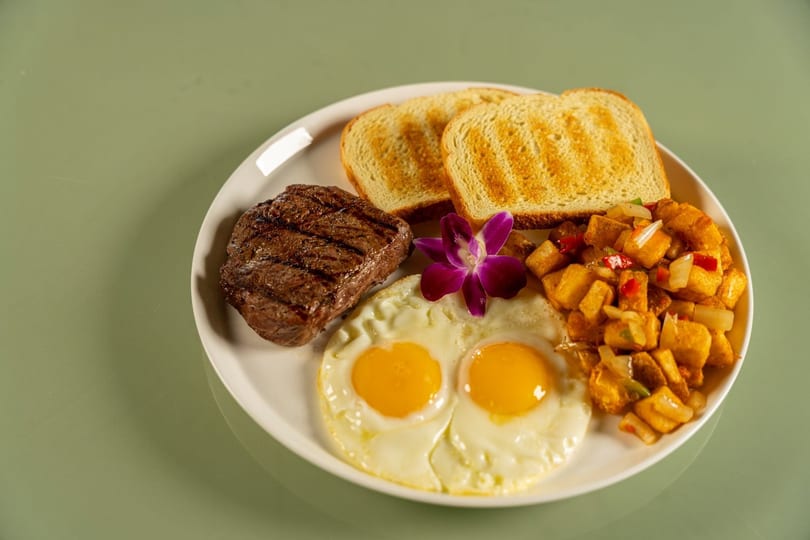 Grilled steak, two fried eggs, toast, and seasoned potatoes garnished with a purple orchid.