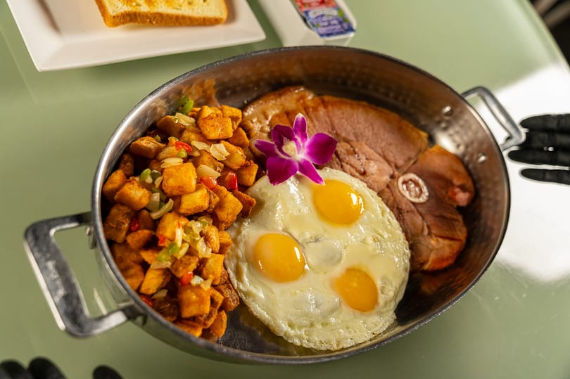 Breakfast skillet with three fried eggs, ham, home fries, and a purple orchid garnish.