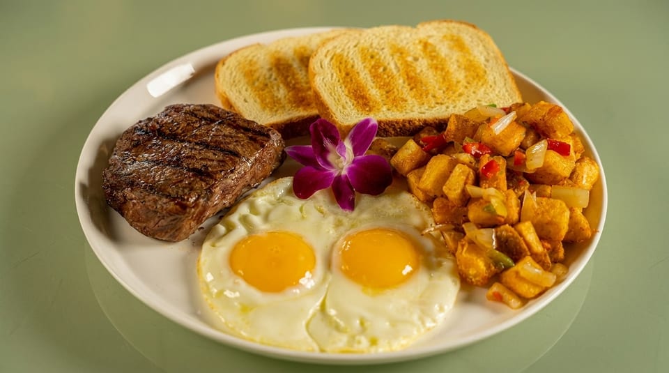 A breakfast plate with grilled steak, two fried eggs, home fries, toast, and a purple orchid.