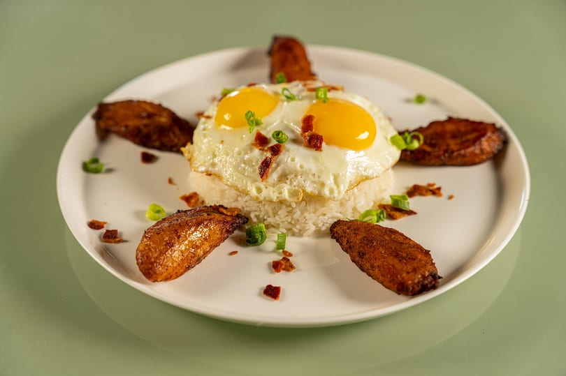 Plate of rice topped with two fried eggs, bacon, green onions, and fried plantains.